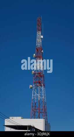 Alto torre di telecomunicazioni rossa e bianca con antenne a parabole contro un cielo blu limpido, edificio sul tetto, Thailandia Foto Stock