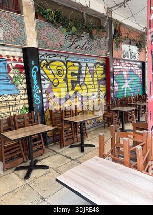 L'esterno del Folia (Nest) Cafe' e' chiuso per affari ad Atene, Grecia. Sedie e tavoli in legno impilati all'esterno del ripostiglio coperto da persiane e ricoperti dal vibrante urb Foto Stock