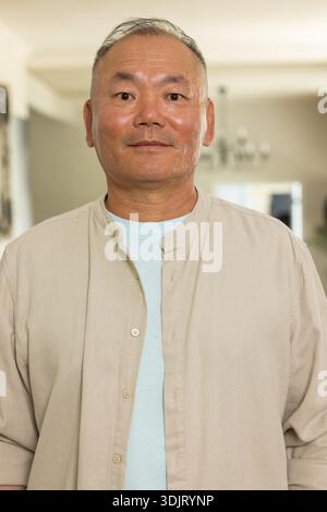Uomo asiatico maturo in piedi a casa con una camicia beige sopra una t-shirt azzurra, un lampadario e un'arte incorniciata Foto Stock