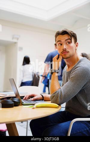 Un giovane premuroso che guarda lontano mentre si siede con un portatile a un tavolo nel campus universitario Foto Stock