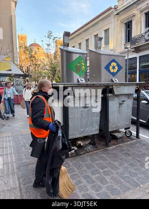 Pulizia stradale di Atene con bidoni di riciclaggio e scopa su ciottoli. Scena urbana frenetica in Grecia. Architettura tradizionale e sfondo della cupola della chiesa. Foto Stock