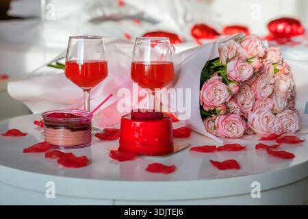 Primo piano di due bicchieri con bevanda rossa, dessert a forma di cuore, rose rosa e petali di rosa su un tavolo. Foto Stock
