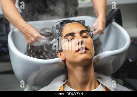 Giovane donna che si rilassa a occhi chiusi mentre le mani del parrucchiere si lavano delicatamente i capelli con shampoo e acqua calda, creando vapore in un salone professionale Foto Stock