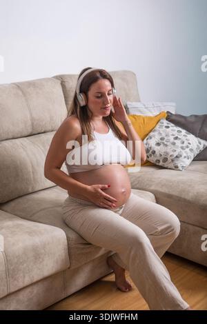 Una donna incinta si siede comodamente sul divano, ascoltando musica dolce tramite le cuffie. Culla dolcemente il suo bambino bump mentre si gode la setta Foto Stock