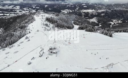 Colpo di drone su una pista da sci innevata tra montagne boscose. Vista aerea ad alto angolo di una stazione sciistica con seggiovie, sciatori, pinete e innevate Foto Stock