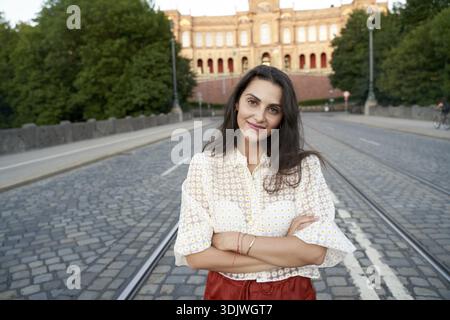 Sorridente con calma e calma, una donna sicura di sé si erge su una strada con le braccia incrociate, indossando una camicia a motivi geometrici, su Maximiliansbruecke davanti a ma Foto Stock