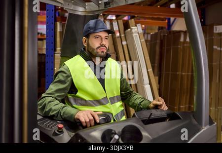 Operatore di carrelli elevatori a forche da magazzino con giubbotto di sicurezza che guida il caricatore tra gli scaffali Foto Stock
