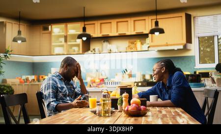 Uomo afroamericano che lavora troppo e donna che chiede una pausa, mostrandogli il segnale di timeout e chiudendo il suo portatile. Fidanzato che lavora da lavoro ignora la sua ragazza durante la colazione. Foto Stock