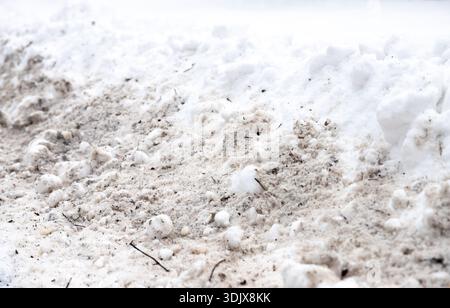 Una pila di neve allinea il bordo di una superficie asfaltata contrassegnata con le tracce degli pneumatici. Il terreno mostra segni di fusione e ricongelamento, con macchie piene e ghiacciate Foto Stock