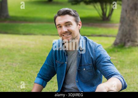 Uomo di mezza età seduto su un prato erboso nel parco, indossa una camicia in denim su una t-shirt grigia e sorride. Adulti, stile di vita, luce solare, stock, all'aperto, natura, c Foto Stock