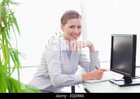 donna degli anni '20 seduta a una scrivania di vetro con indosso una camicia blu, che tiene la penna sopra i documenti del monitor, dell'impianto. Professionale, ufficio, Highkey, fogliame, aziendale, f Foto Stock