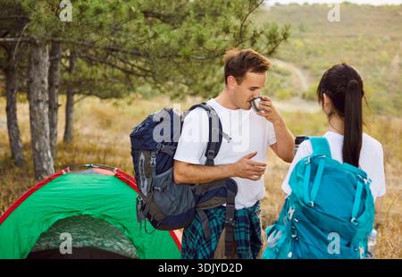 Un paio di turisti in campeggio con zaini vicino alla tenda della foresta, uomo che beve dalla tazza Foto Stock