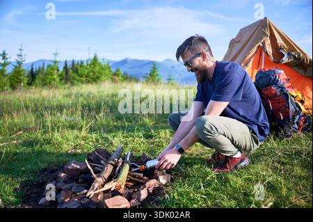 Uomo barbuto con occhiali da sole e abbigliamento casual all'aperto che prepara il fuoco nel panoramico prato di montagna. Tenda arancione e zaino sullo sfondo, con vegetazione lussureggiante e montagne lontane sotto il cielo azzurro. Foto Stock