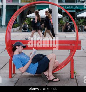 Un giovane uomo rilassa la lettura su una panchina rossa circolare nel locale pubblico urbano di London South Bank mentre i pedoni passano davanti. Foto Stock