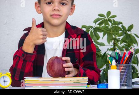 Scolaro che mostra i pollici in alto e tiene una mela alla sua scrivania di studio. Foto Stock