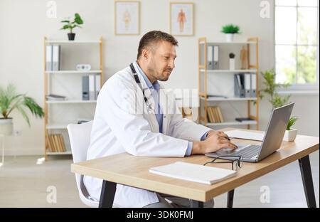 Maschio medico è seduto alla sua scrivania in ospedale e lavorare su computer portatile Foto Stock