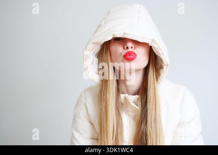 Donna sorridente con rossetto rosso che indossa una giacca invernale bianca e un cappuccio su sfondo chiaro Foto Stock