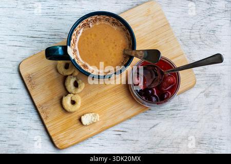 Vista dall'alto di una tazza di caffè su un asse di legno con biscotti, burro e un vaso aperto di marmellata di mirtilli con un cucchiaio, su un tavolo leggero con spazio per le copie. Foto Stock