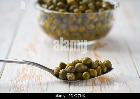 Capperi marinati in un cucchiaio di metallo su un tavolo di legno bianco, messa a fuoco selettiva. Capperi commestibili pronti da mangiare. Foto Stock