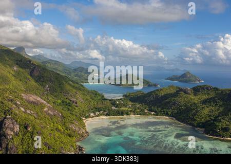 Vista aerea di dove le acque turchesi si incontrano con lussureggianti colline verdi sotto un cielo disseminato di morbide nuvole, Seychelles e Seychelles. Foto Stock