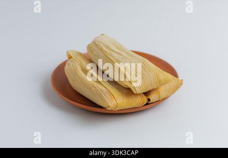 Dettaglio dall'alto verso il basso dei tradizionali tamales messicani su un piatto di argilla arancione, evidenziando la consistenza della buccia di mais secca. Foto Stock