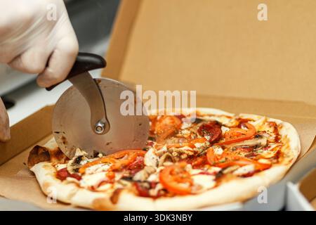 Lo chef taglia la pizza a base di carne calda con un taglierino a ruota tonda nella scatola di consegna. Affettare il processo alimentare da asporto. Foto Stock