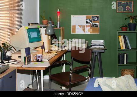 Area di lavoro vintage per home Office con monitor retro per computer, tastiera, mouse, telefono, stampante e varie forniture per ufficio disposte su una scrivania in legno contro una parete verde con bacheca Foto Stock