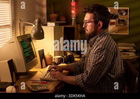 Uomo di mezza età caucasico con barba seduto alla scrivania a scrivere sul monitor di un computer d'epoca in un ufficio domestico, circondato da tecnologia retrò e forniture per ufficio, focalizzato sullo schermo Foto Stock