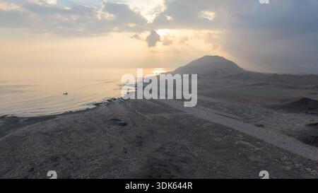 Vista aerea dell'aspra costa vulcanica al tramonto con mare calmo, raggi del sole che si infrangono attraverso le nuvole e il paesaggio di montagna arido con una luce dorata soffusa Foto Stock