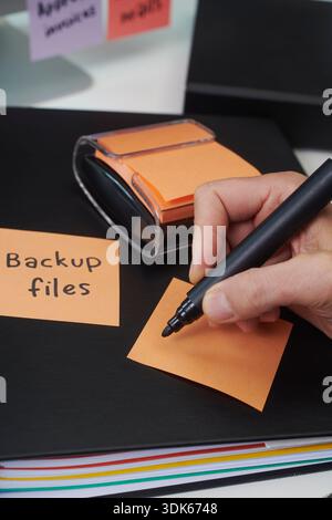una stretta visuale di una nota adesiva arancione che sta per essere scritta da un uomo con un pennarello nero vicino a un distributore di appunti, che mostra i file di backup del testo promemoria o Foto Stock