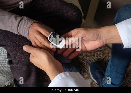 durante una visita a casa, un operatore sanitario misura la saturazione di ossigeno mentre una donna anziana siede comodamente su una poltrona Foto Stock