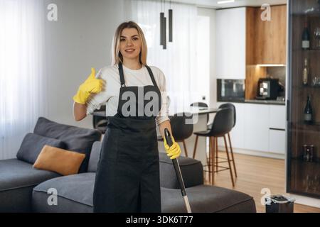 Con il pollice in alto, in piedi. Una donna lavoratrice di pulizie professionale è una stanza domestica. Foto Stock