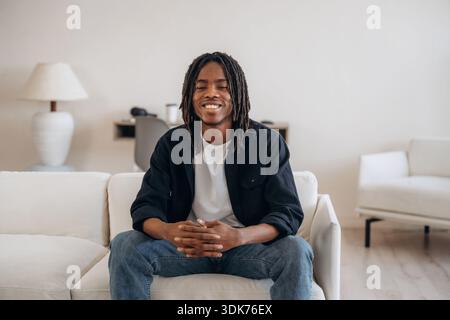 L'allegro ragazzo afroamericano è seduto sul divano a casa e sorride. Foto Stock