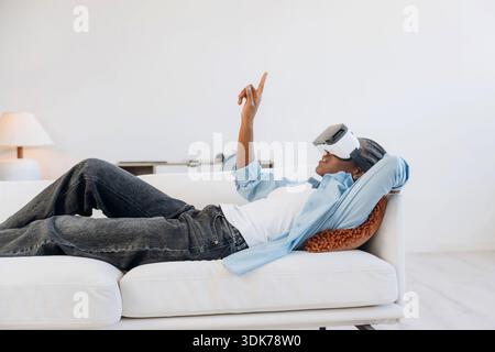 Vista laterale, sdraiato e indossato il visore VR. La donna è a casa in una stanza domestica. Foto Stock