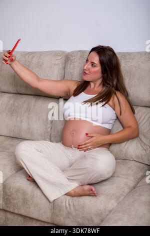 Una donna allegra si siede comodamente su un divano morbido, toccando delicatamente il suo seno del bambino e catturando la sua felicità con un selfie. La luce del sole riempie delicatamente il Foto Stock