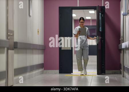 Giovane donna adulta che accede al corridoio del centro vascolare riesaminando la cartella paziente Foto Stock