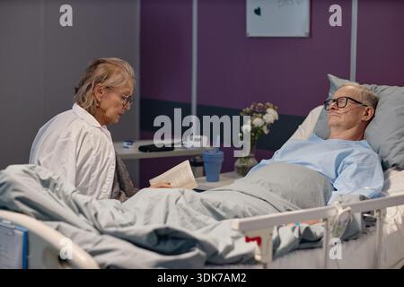Donna anziana che legge Libro per l'uomo anziano che riposa in ospedale Foto Stock