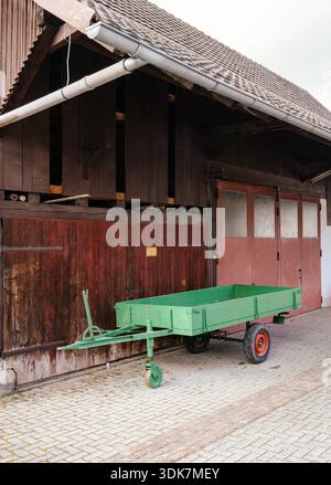 Rustico rimorchio agricolo verde parcheggiato davanti alle porte del fienile di legno su un cortile di ciottoli sotto il tetto piastrellato che trasporta il patrimonio agricolo rurale Foto Stock