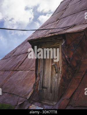 Vista di una porta di legno intemprata all'interno dell'abbraccio arrugginito e testurizzato di un tetto spiovente sullo sfondo di un cielo nuvoloso, Lukomir, Bosn Foto Stock