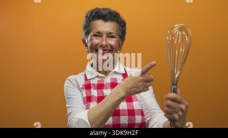 Donna anziana sorridente con capelli grigi che reggono la frusta e indossa grembiule rosso su sfondo arancione. Foto Stock