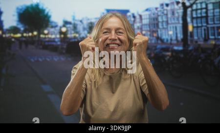 Donna anziana che stringe le mani al petto in una strada accanto agli edifici del canale e parcheggia le biciclette, sorridendo con i pugni alzati per festeggiare; gioia resilienza. Foto Stock