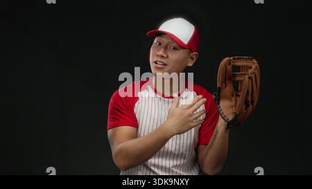 Il giovane cinese in uniforme da baseball posa con sicurezza con il guanto su sfondo nero isolato. Foto Stock