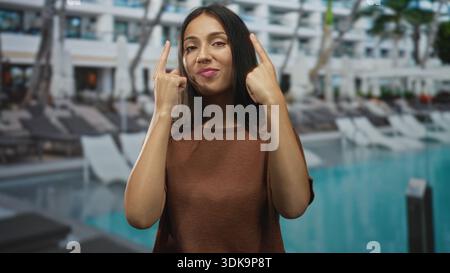 Donna ispanica punta entrambe le dita indice verso l'alto sui lettini della piscina all'aperto dell'hotel mentre sorride; fiducia giocosa. Foto Stock