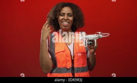 Donna che tiene in mano un drone e fa una pompa a pugno con mano visibile mentre sorride in studio con sfondo rosso; risultato emozionante. Foto Stock