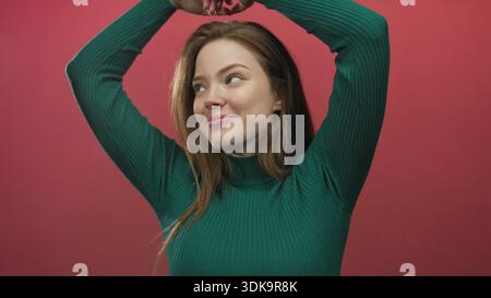 Donna sorridente che forma una forma a cuore con le mani in un ambiente da studio rosso, guardando direttamente la fotocamera con le braccia alzate; amore. Foto Stock