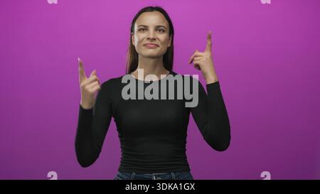 La donna punta il dito indice verso l'alto nello studio magenta mentre guarda in alto e indossa camicia e jeans neri; curiosità. Foto Stock