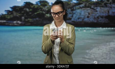 Donna con occhiali da vista, utilizza lo smartphone tenendo in mano il dispositivo e guardando in basso in uno studio con sfondo da spiaggia; concentrazione. Foto Stock