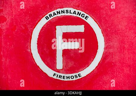 Una parete dipinta di rosso mostra un cerchio bianco con la lettera F e la scritta bilingue della manichetta antincendio, che segna chiaramente la stazione dei manichette antincendio per una rapida emergenza Foto Stock