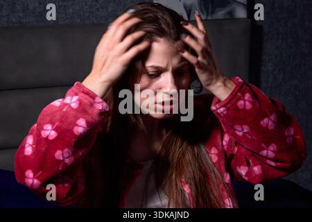 Questa immagine cattura una giovane donna in un momento di intenso disagio emotivo. Seduto a letto e rivestito in un'accogliente veste rossa adornata da un allegro trifoglio Foto Stock