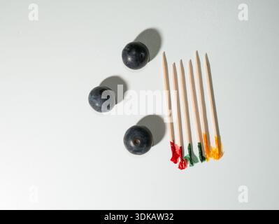 Un layout minimalista con tre mirtilli freschi disposti accanto a cinque stuzzichini da cocktail in bambù con punte colorate e fritte su un semplice suon bianco Foto Stock